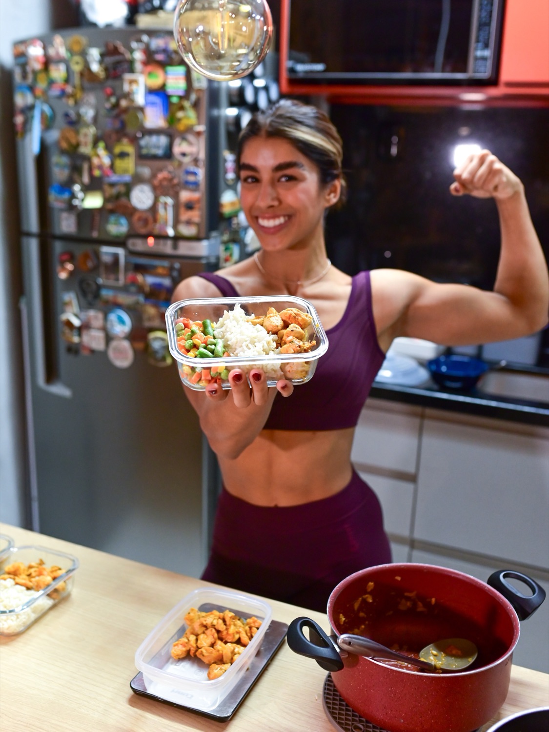 Kakau Farias segurando uma marmita montada e flexionando o braço na cozinha